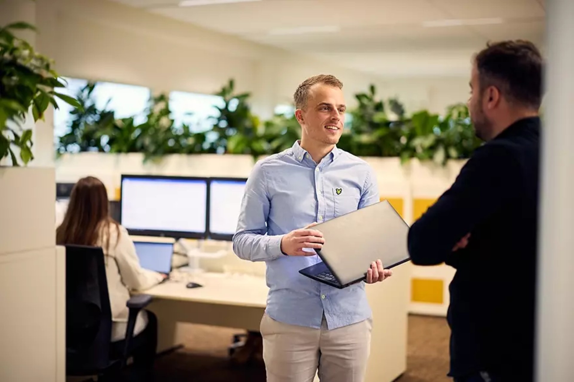 Twee mannen praten in een modern kantoor, terwijl een vrouw aan een bureau werkt.